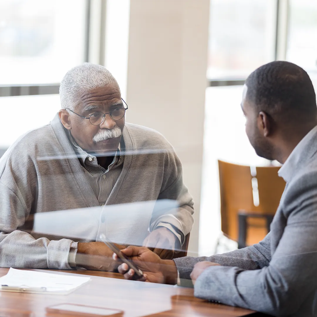 A senior male customer sits at a desk, listening to a male employee who is holding a mobile phone.