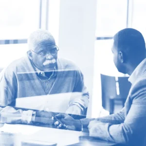 A senior male customer sits at a desk, listening to a male employee who is holding a mobile phone.