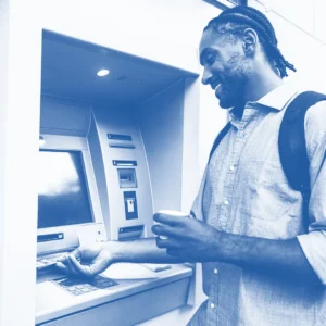 Pictured is a man wearing a blue, button-down shirt and backpack retrieving cash from an ATM. The man is smiling and holding a coffee, evoking the ease of the process.