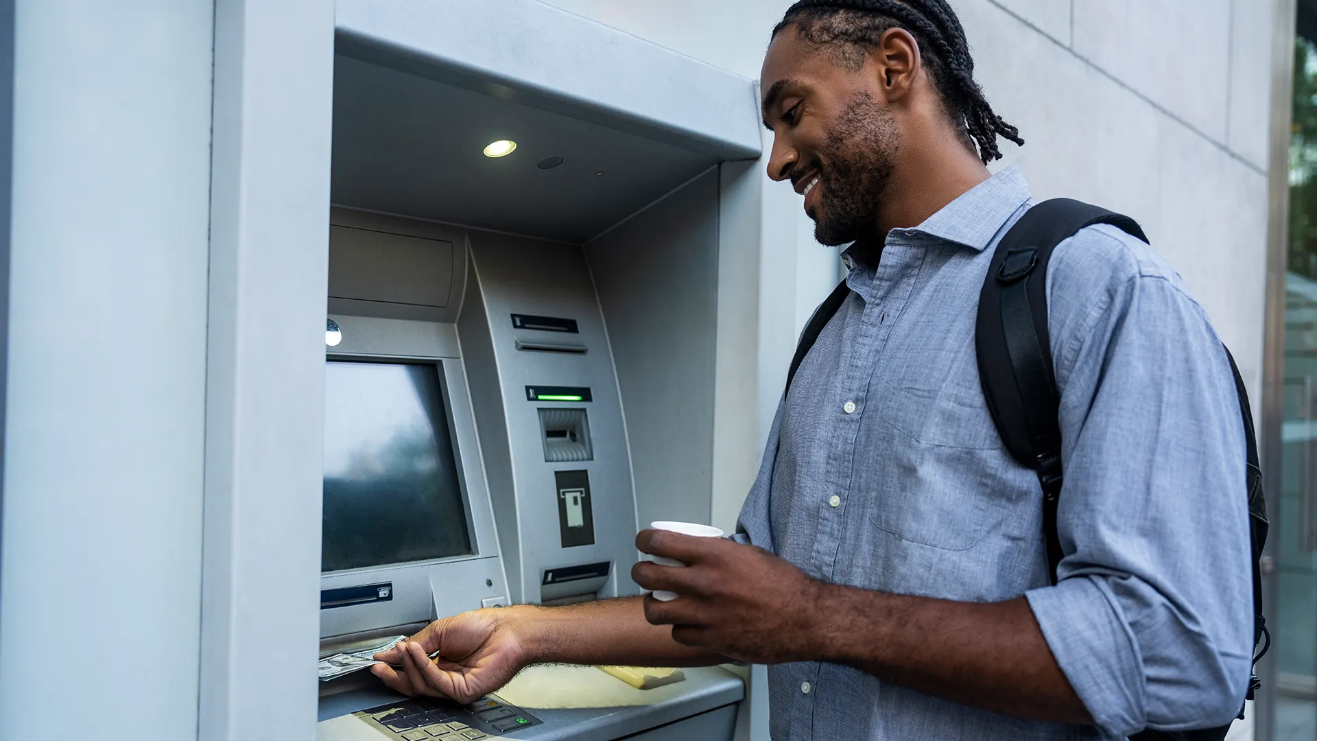 Pictured is a man wearing a blue, button-down shirt and backpack retrieving cash from an ATM. The man is smiling and holding a coffee, evoking the ease of the process.